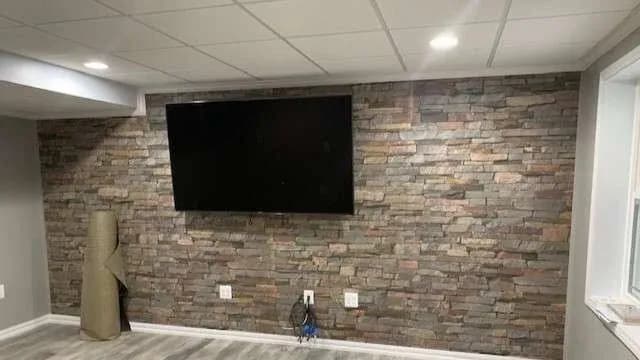 Basement room with a stone accent wall featuring a large TV. A rolled-up rug leans against the left wall. The space is modern and neatly organized.