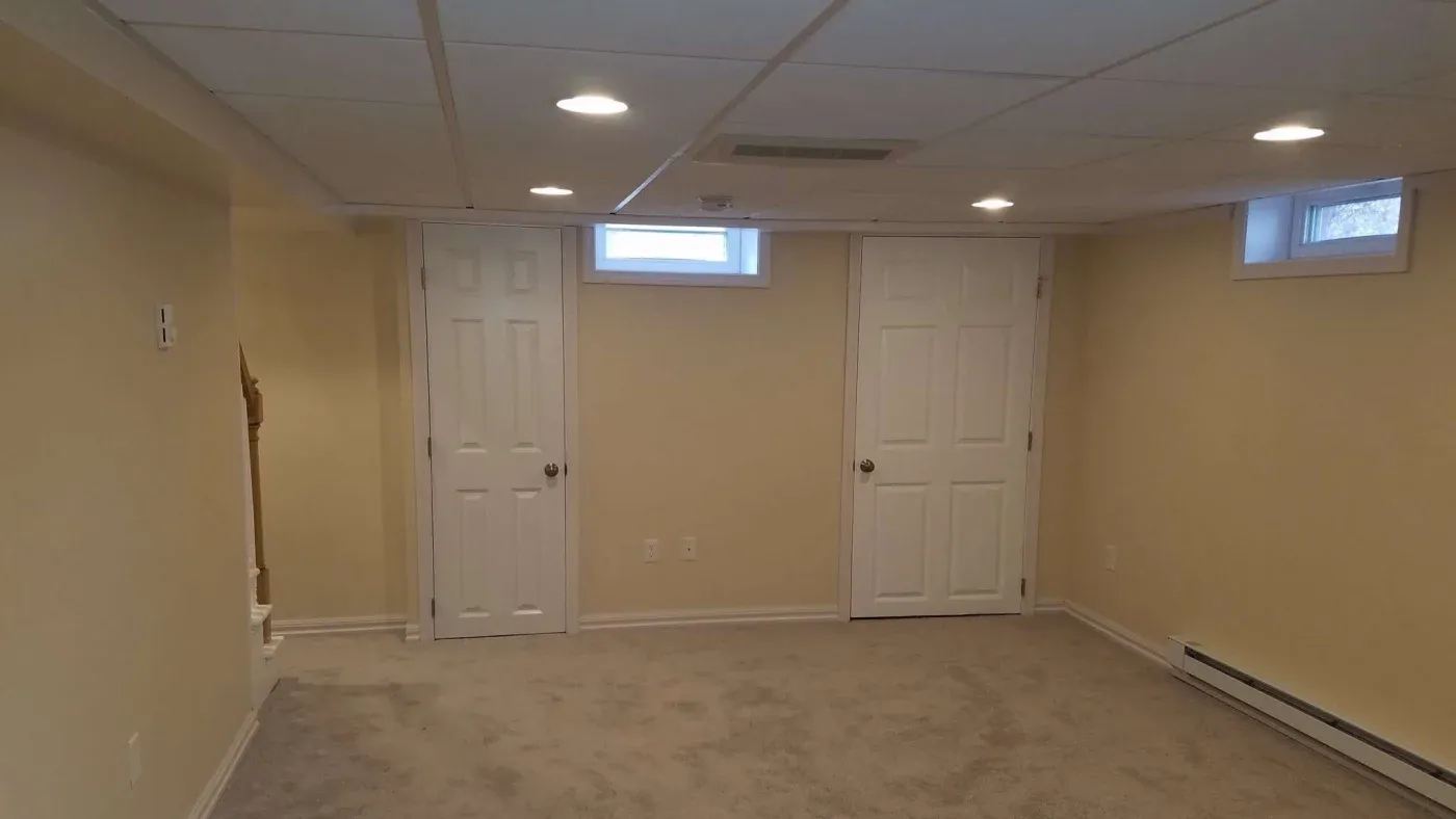 Spacious basement room with beige walls and carpet. Two white doors and three small windows; bright ceiling lights create a warm, inviting atmosphere.