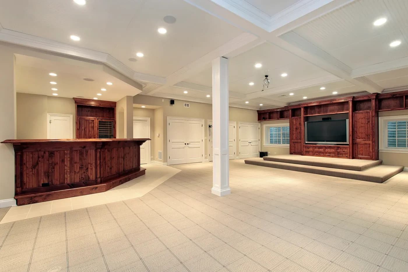 Spacious basement with recessed lighting, featuring a wooden bar and a media area with a large TV on a carpeted platform. Neutral tones create a cozy ambiance.
