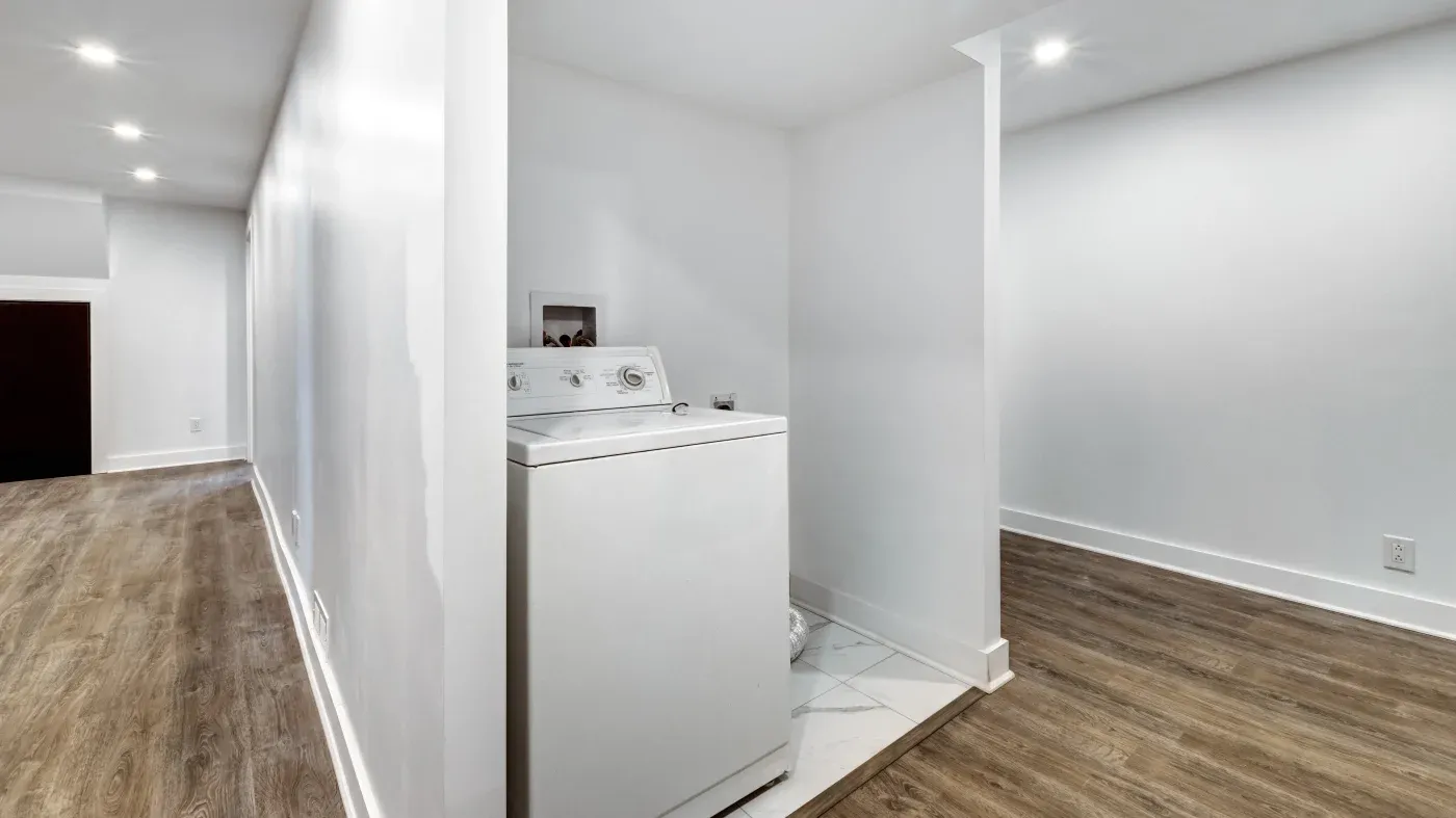 A white washing machine is in a small laundry nook with tiled flooring. The area is bright and minimalistic, connecting to a hallway with wood flooring.