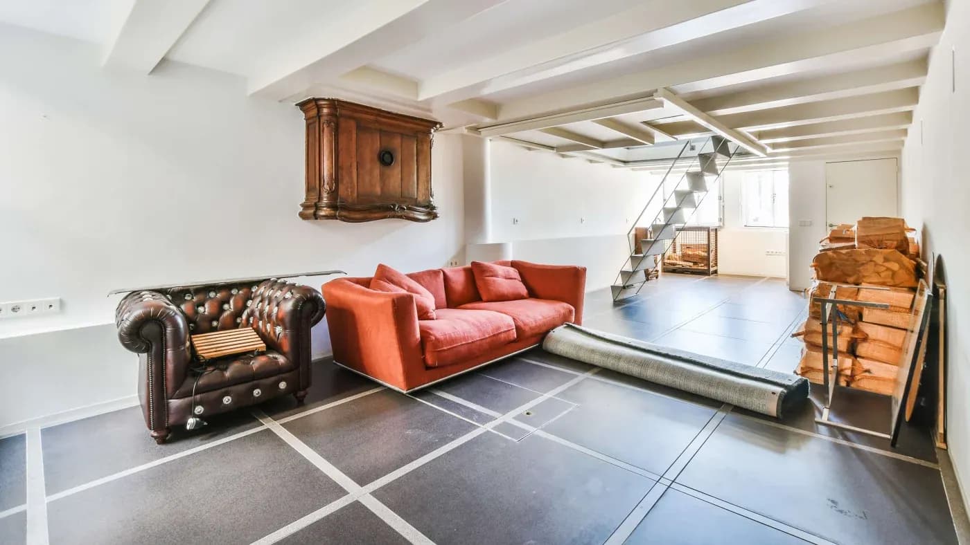 Spacious modern living room with a red couch, brown armchair, wooden chest on the wall, and metal stairs. A cozy, minimalist ambiance.
