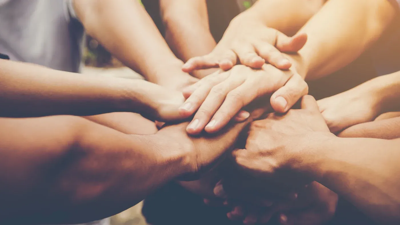 A group of diverse hands stacked together in a display of teamwork and unity. The warm lighting conveys a sense of collaboration and solidarity.