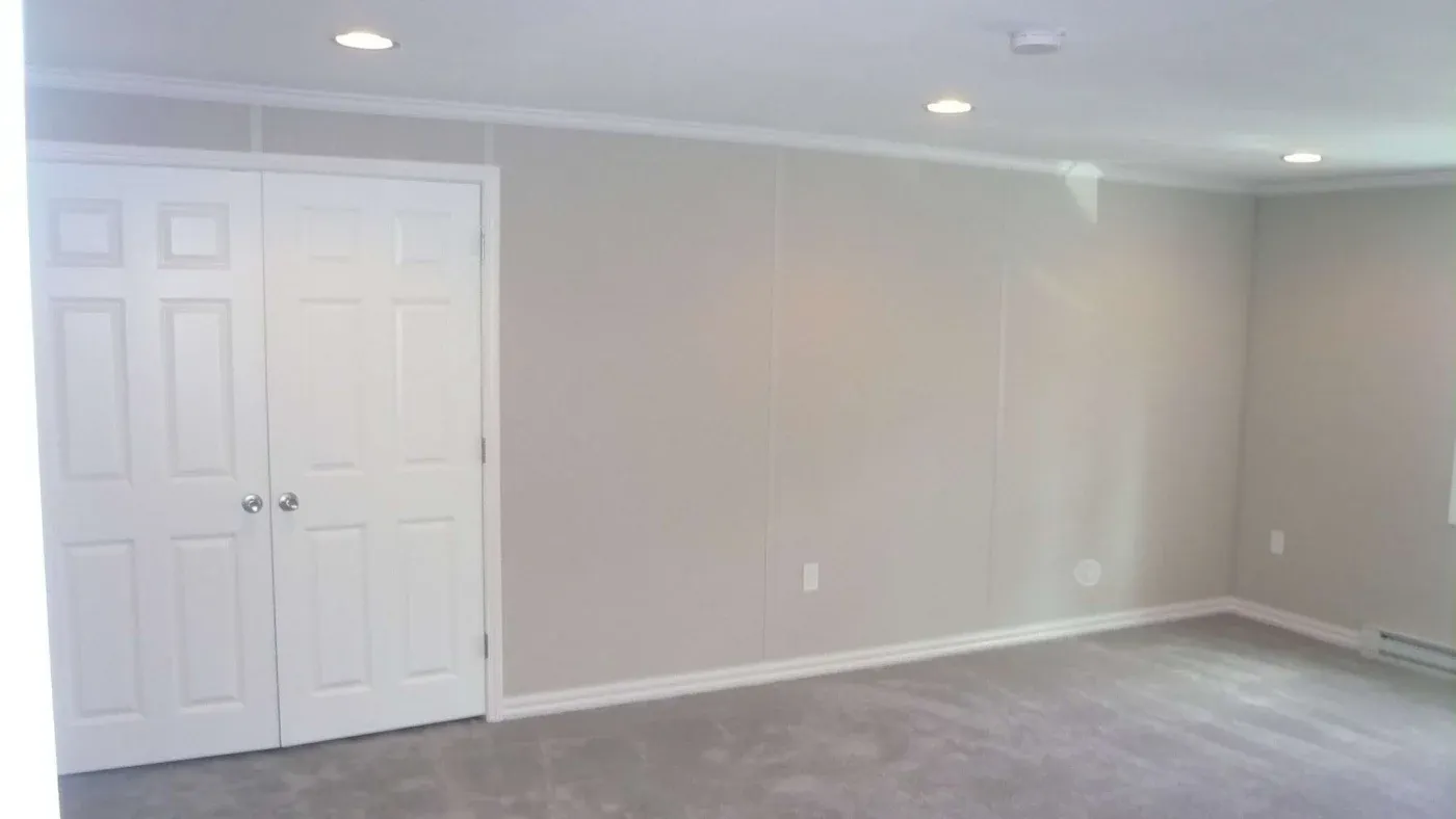 A simple, empty room with beige walls and gray carpet. It features recessed lighting and a white double door. The atmosphere is clean and minimalistic.