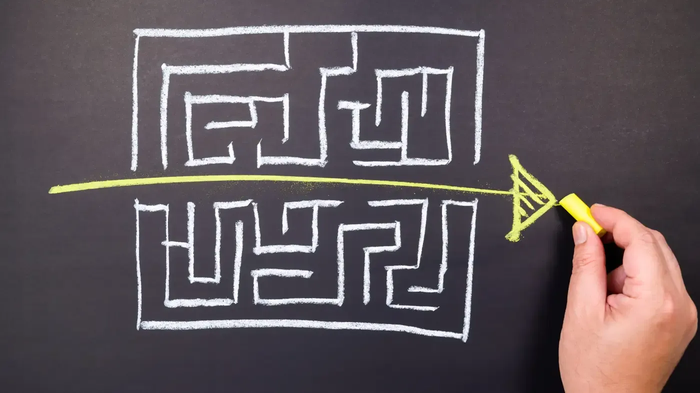 A hand draws a yellow arrow cutting through a white chalk maze on a blackboard, symbolizing finding a straightforward solution through complexity.