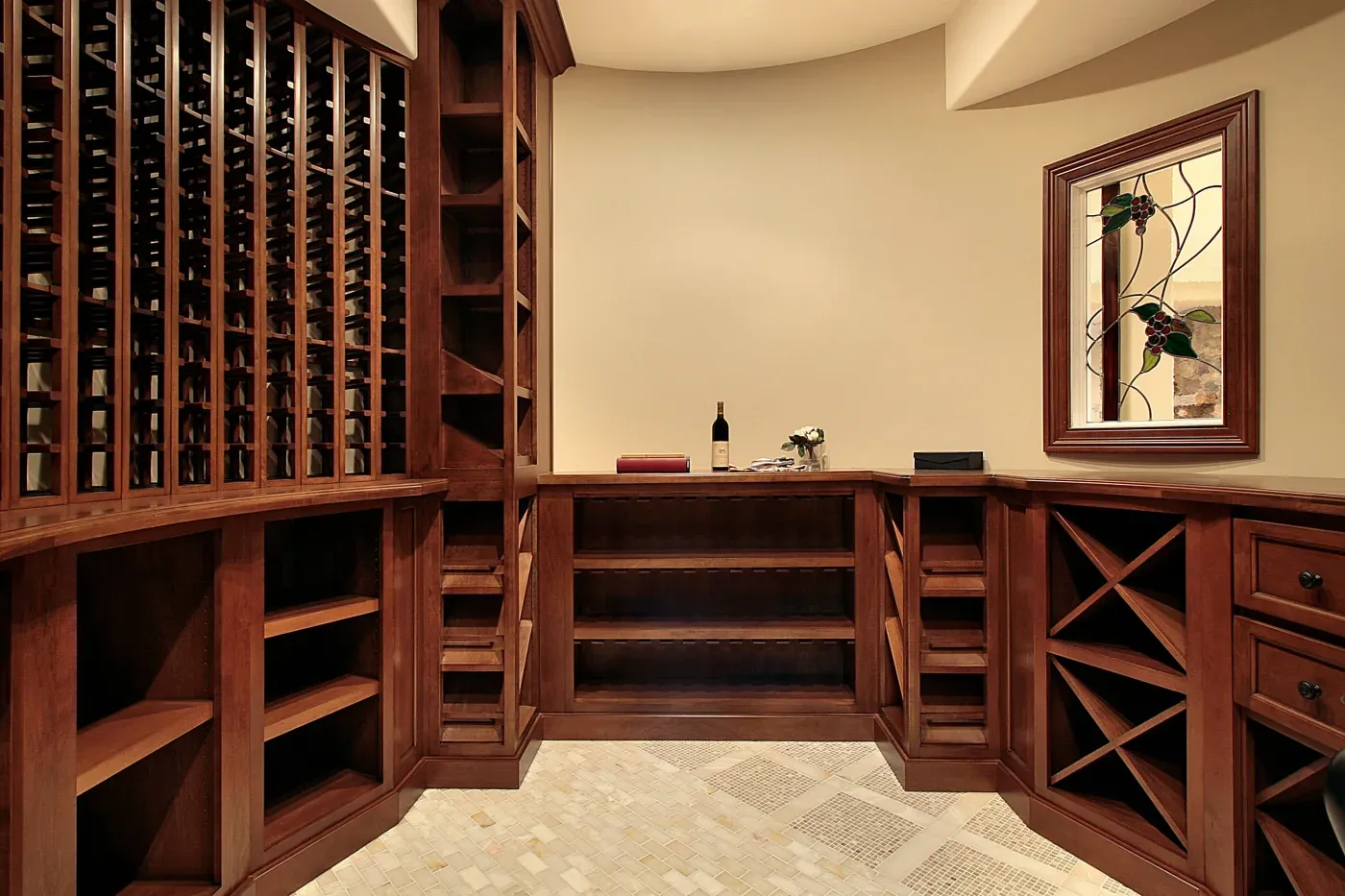 A wine cellar with rich wooden racks, an empty shelf, and a countertop holding a wine bottle. A window with grapevine design adds elegance.