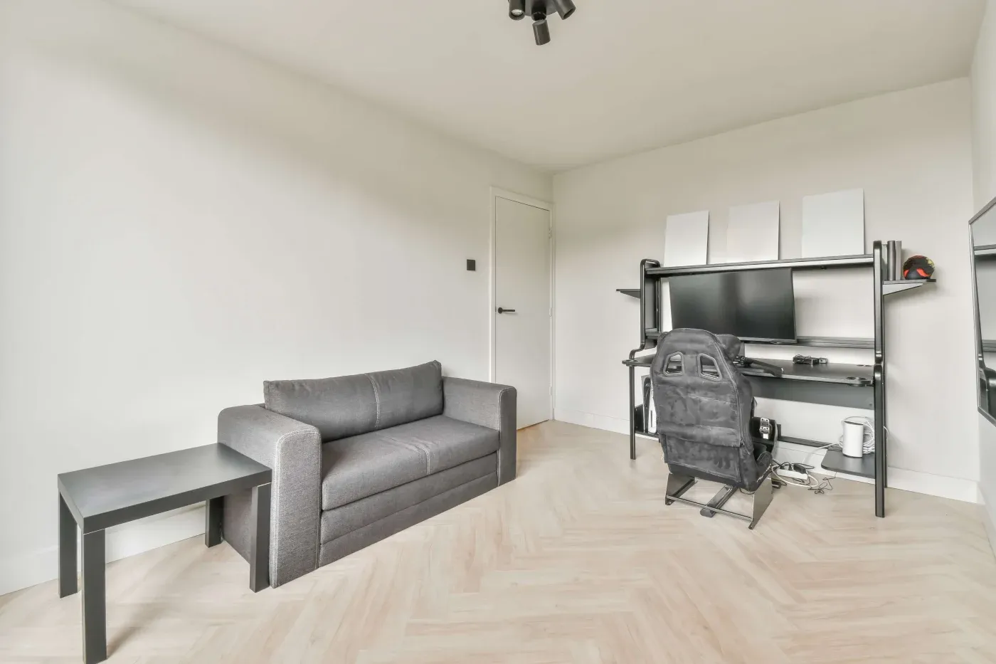 A minimalist room with a gray sofa, black side table, and modern gaming setup. The desk features a computer monitor and gaming chair, creating a sleek ambiance.
