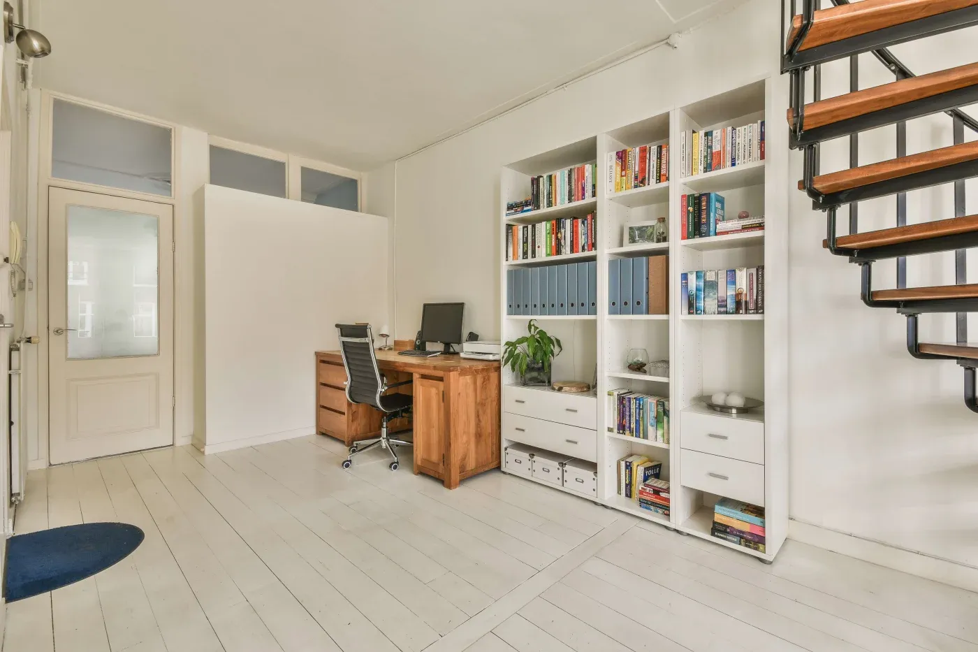 Modern study room with white walls, a wooden desk and chair, a computer, bookshelves filled with books, and a spiral staircase. Bright and organized.