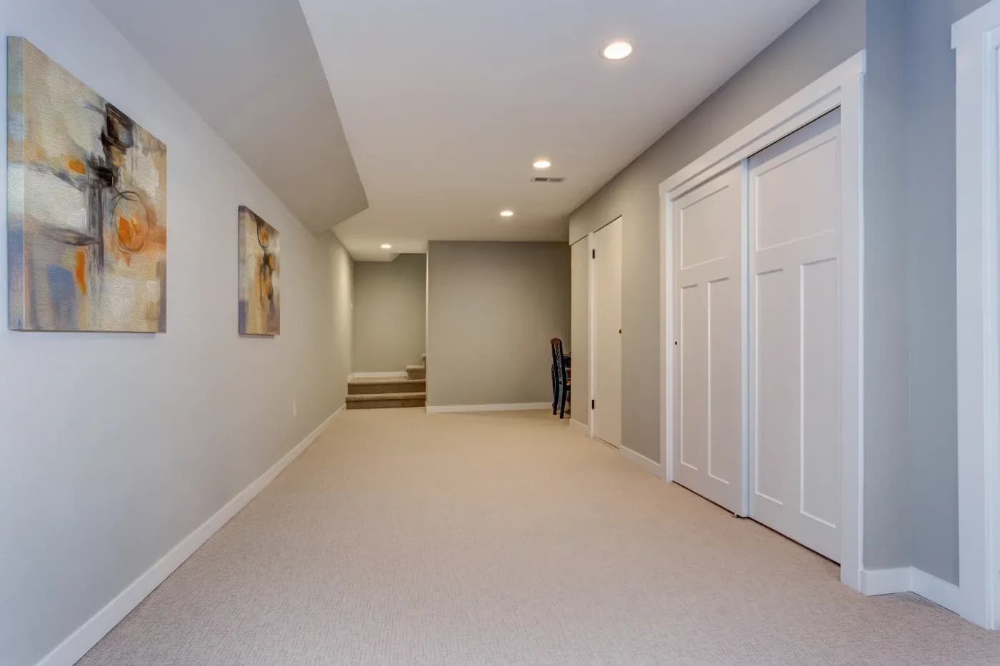 A spacious, empty hallway with beige carpet and gray walls. There are abstract paintings on the left wall and white double doors on the right. A staircase is visible at the end.
