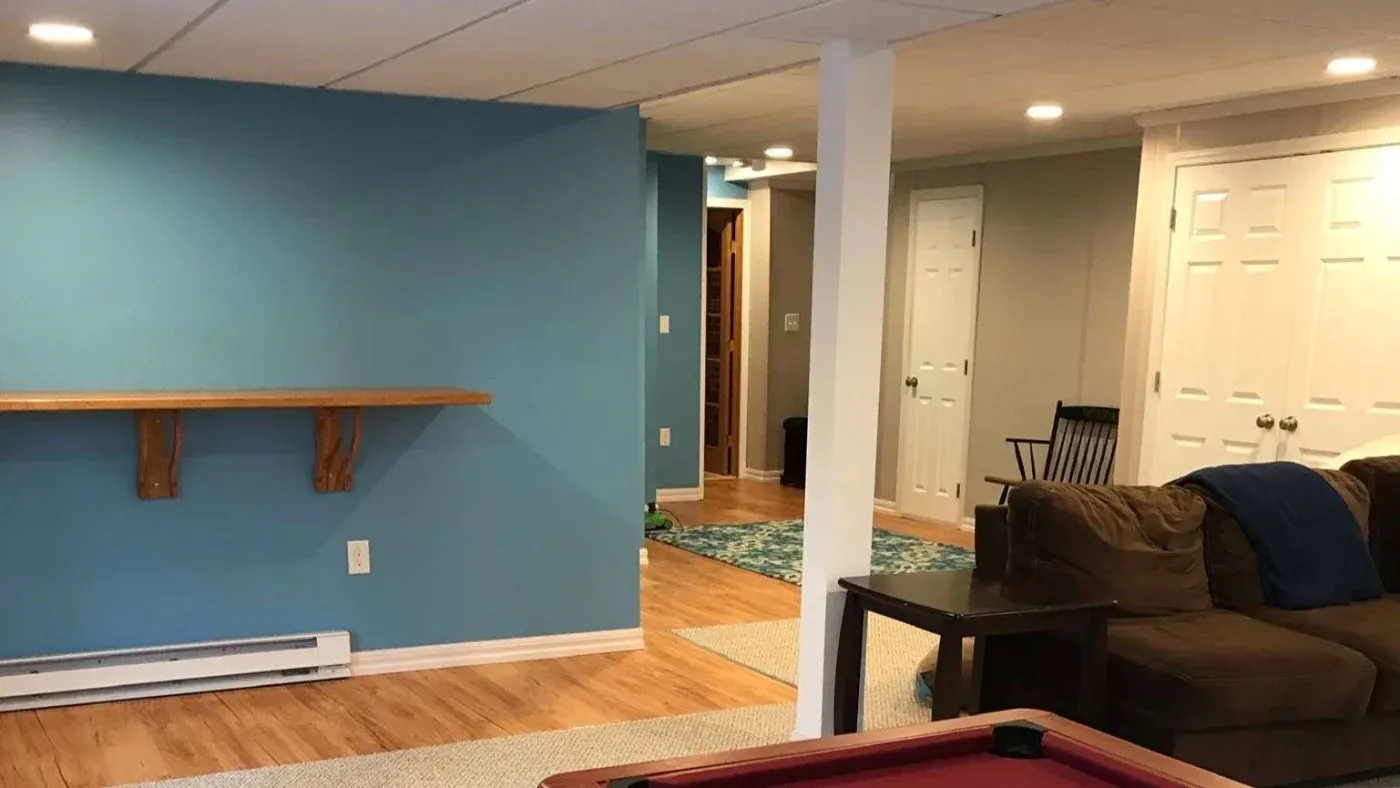 A cozy basement living area with light blue and beige walls, featuring a wall-mounted wooden shelf, brown sectional sofa, and a pool table.