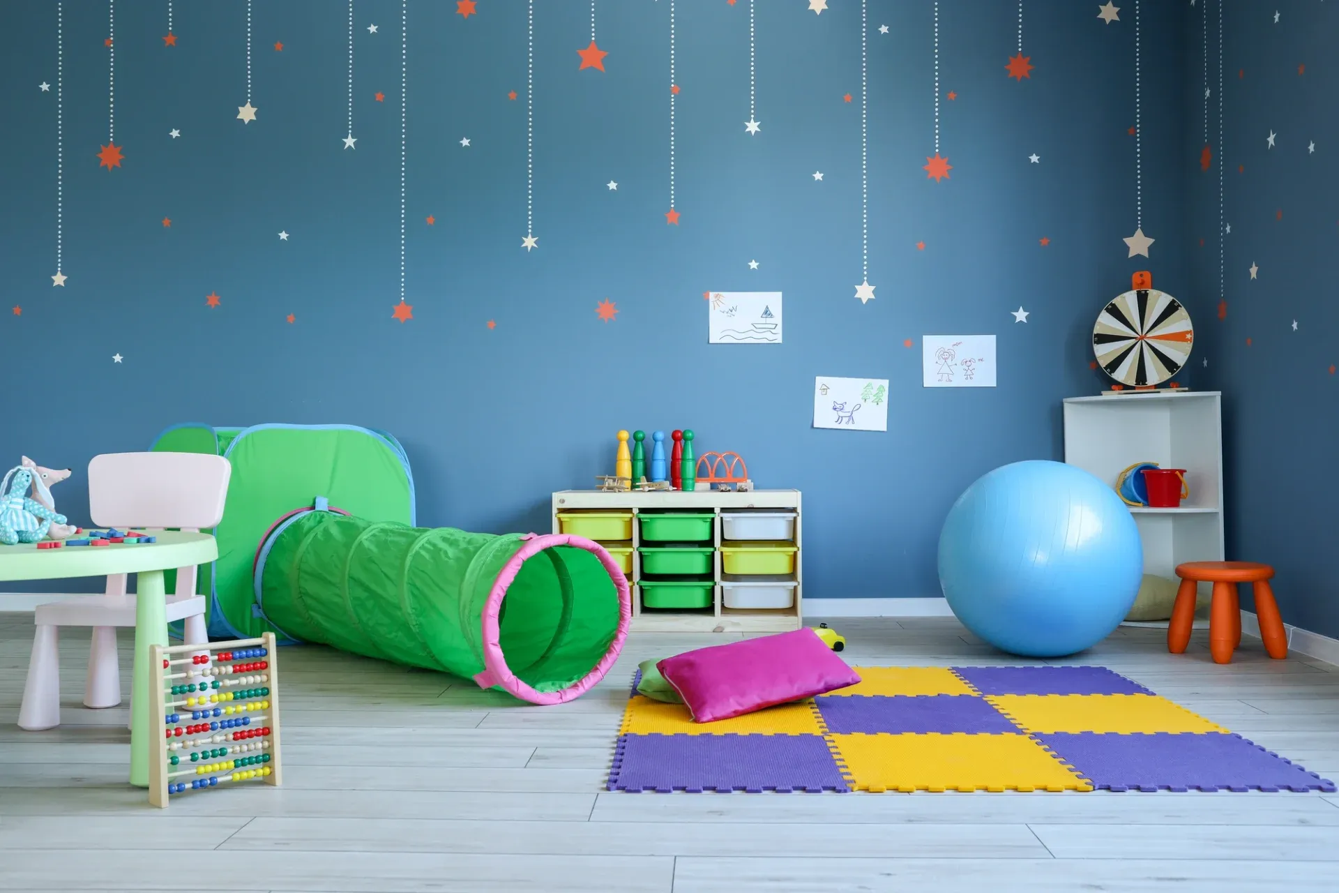A colorful playroom with a blue wall adorned with stars. Features a green tunnel, foam mats, toys, a hop ball, and child-sized furniture, creating a fun, playful atmosphere.