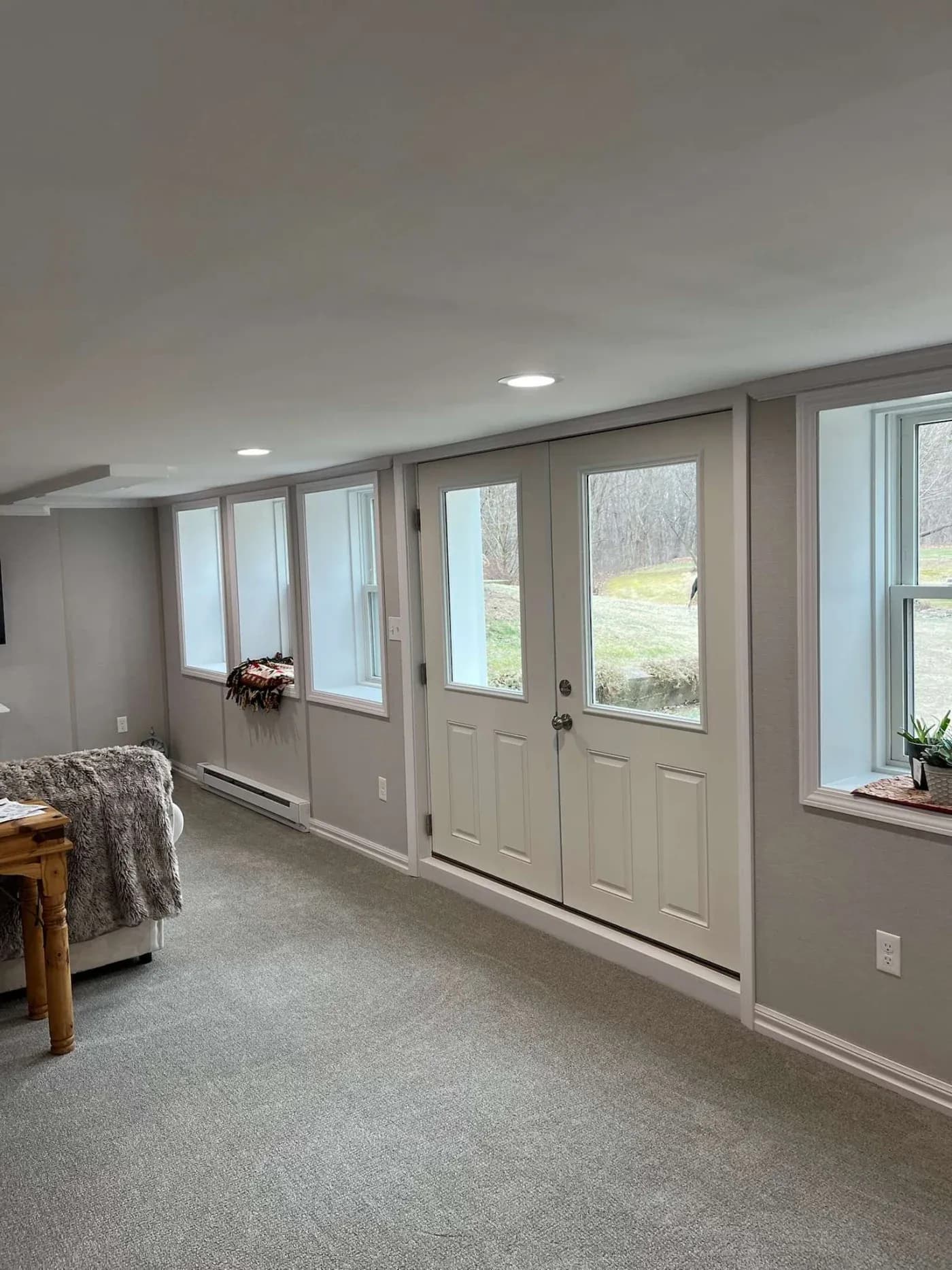 Bright basement room with light gray walls, featuring a row of large windows and glass double doors. Natural light streams in, creating an inviting atmosphere.