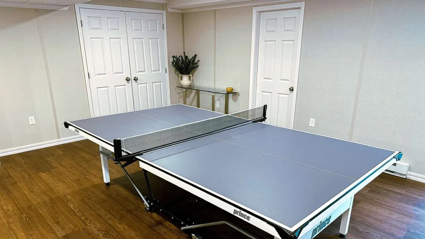 A ping pong table with a net is set up in a room with wooden floors. There are two closed white doors and a table with a plant in the background.