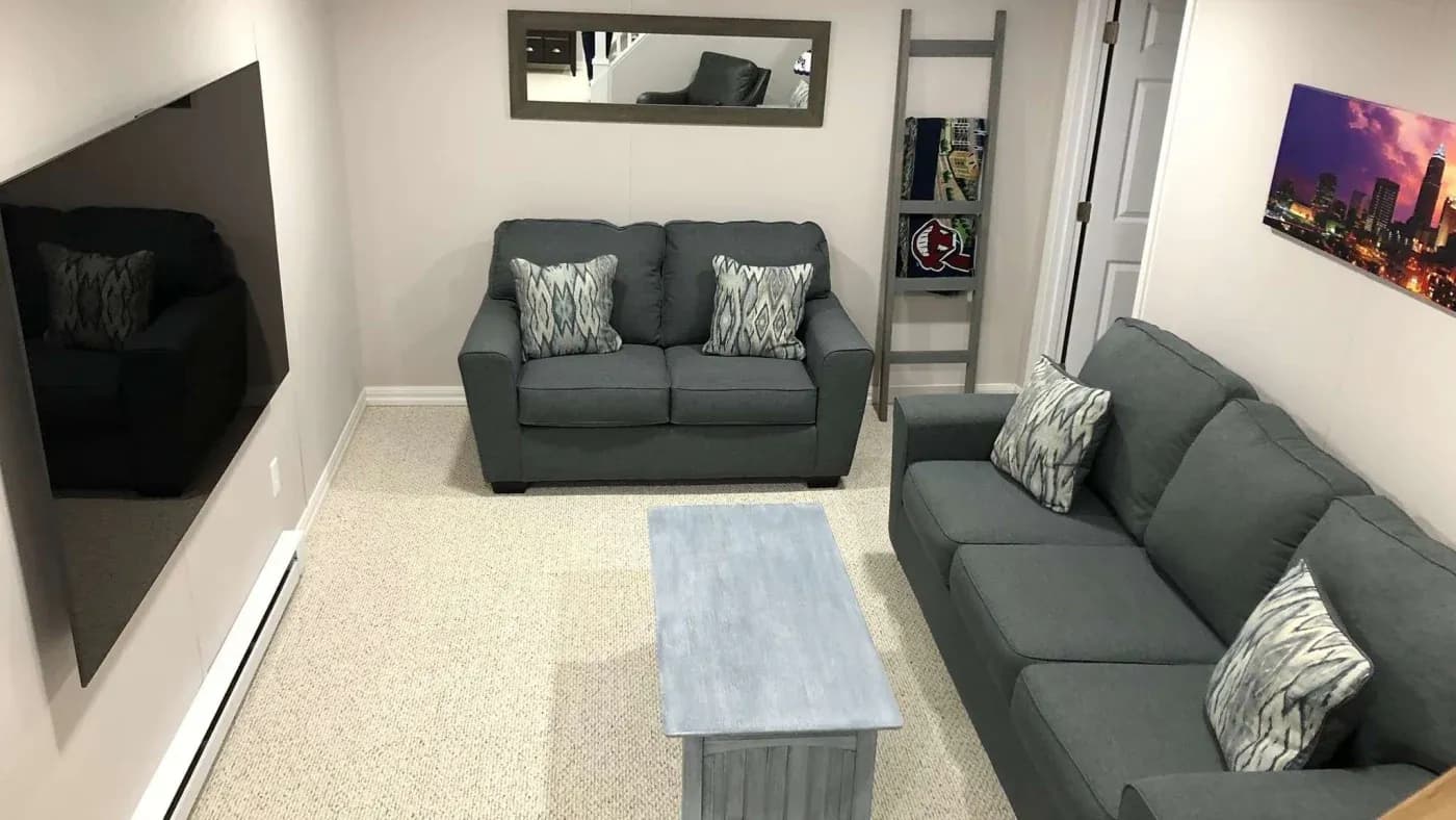 A cozy living room with two gray sofas facing a large wall-mounted TV. A light blue coffee table sits between the sofas. The room has a soft, neutral color palette with patterned throw pillows. A landscape cityscape photo hangs on the wall, and a ladder shelf holds books and decor.