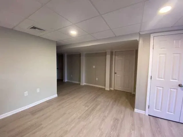 Empty basement room with light wood flooring, beige walls, and a white drop ceiling. Two closed white doors and recessed lighting create a clean, spacious feel.