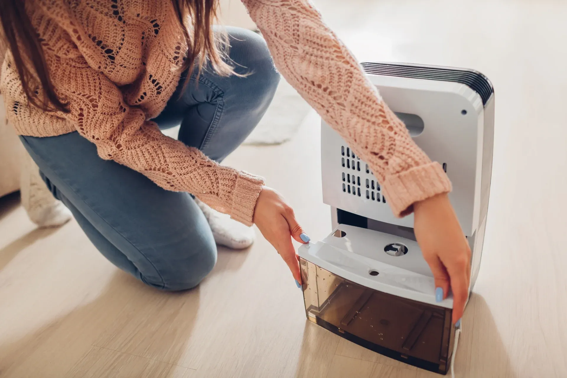 A person in a pink sweater and jeans is kneeling, removing the water tank from a dehumidifier on a wooden floor. The scene suggests maintenance.
