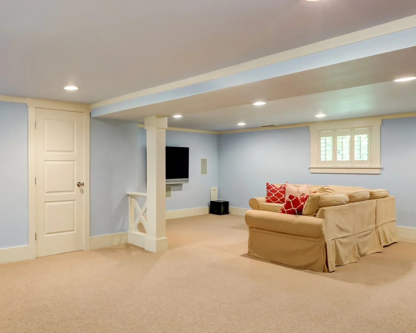 A cozy basement room with light blue walls and beige carpet, featuring a tan sectional sofa with red pillows, a wall-mounted TV, and recessed lighting.