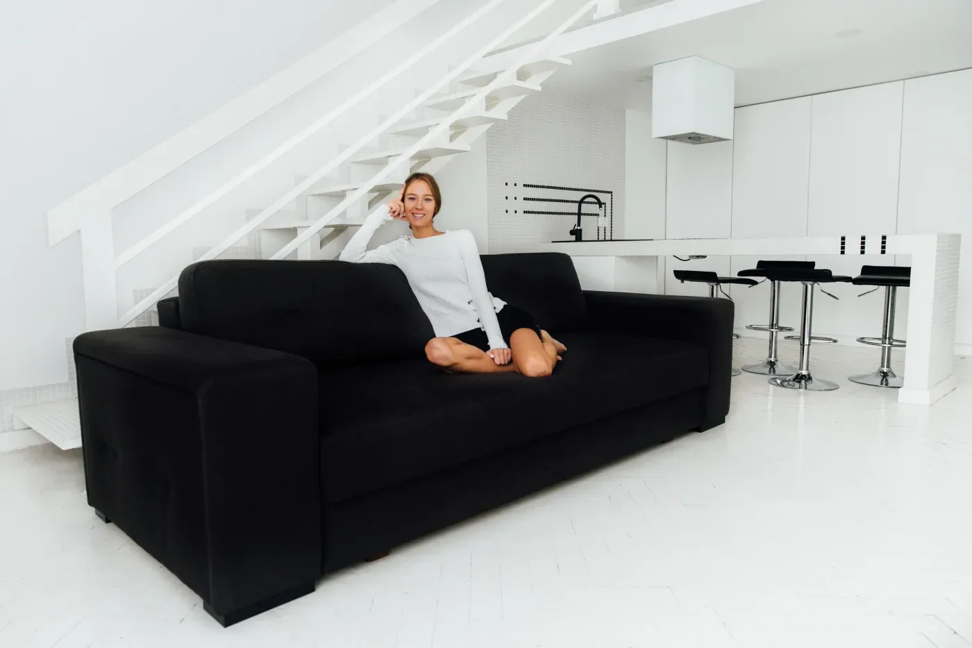 A woman sits relaxed on a black sofa in a modern, white minimalist living space, with a sleek staircase and kitchen area visible in the background.
