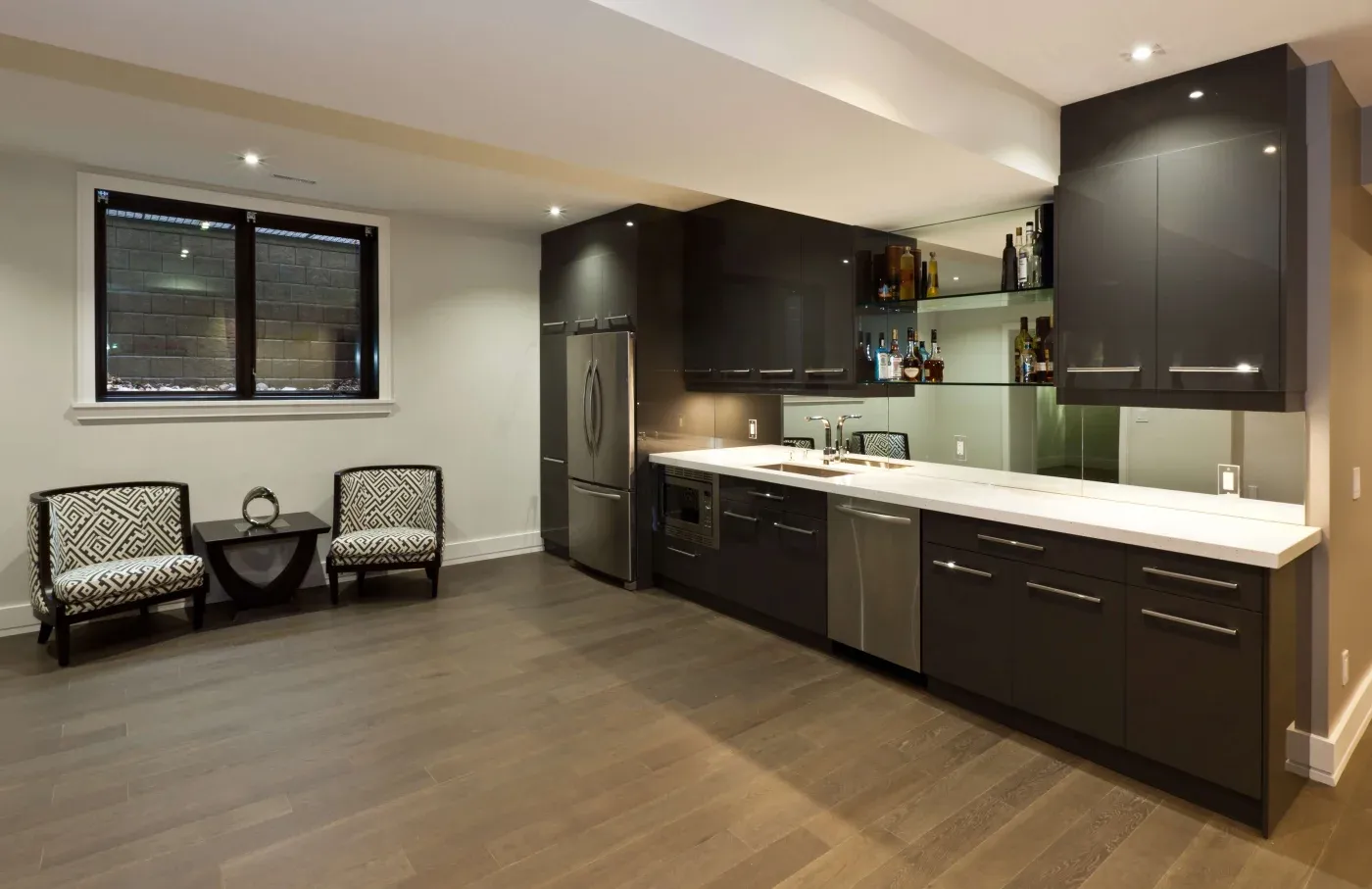 Modern kitchen featuring sleek dark gray cabinets, a stainless steel fridge, and minimalistic decor. Two patterned chairs sit against a light wall.