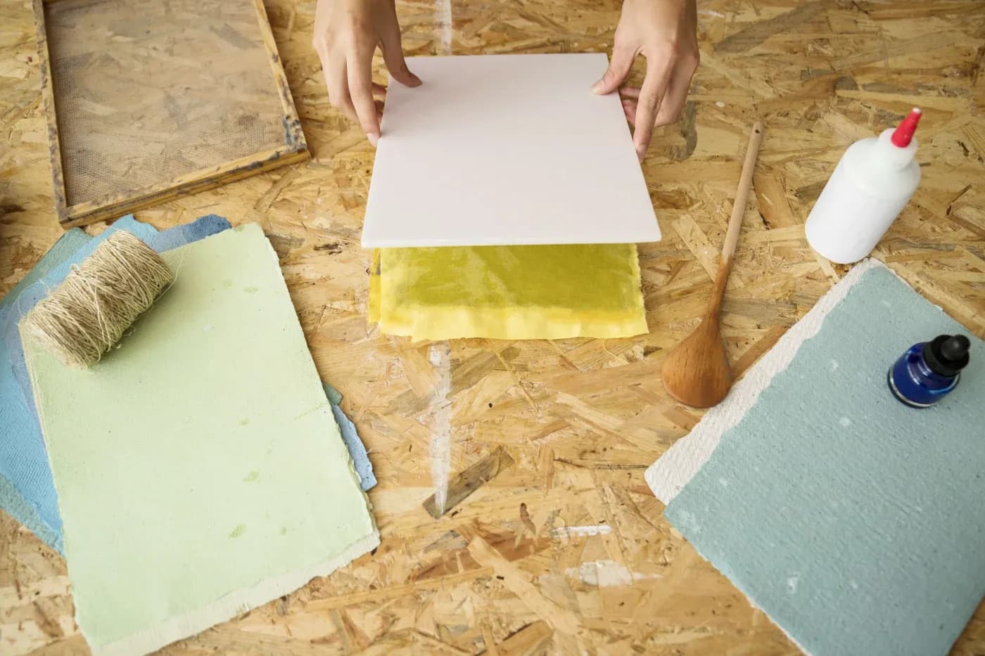 Hands placing a white sheet over yellow paper on a wooden surface. Nearby are colored paper sheets, twine, a wooden spoon, glue, and ink, suggesting a crafting setup.