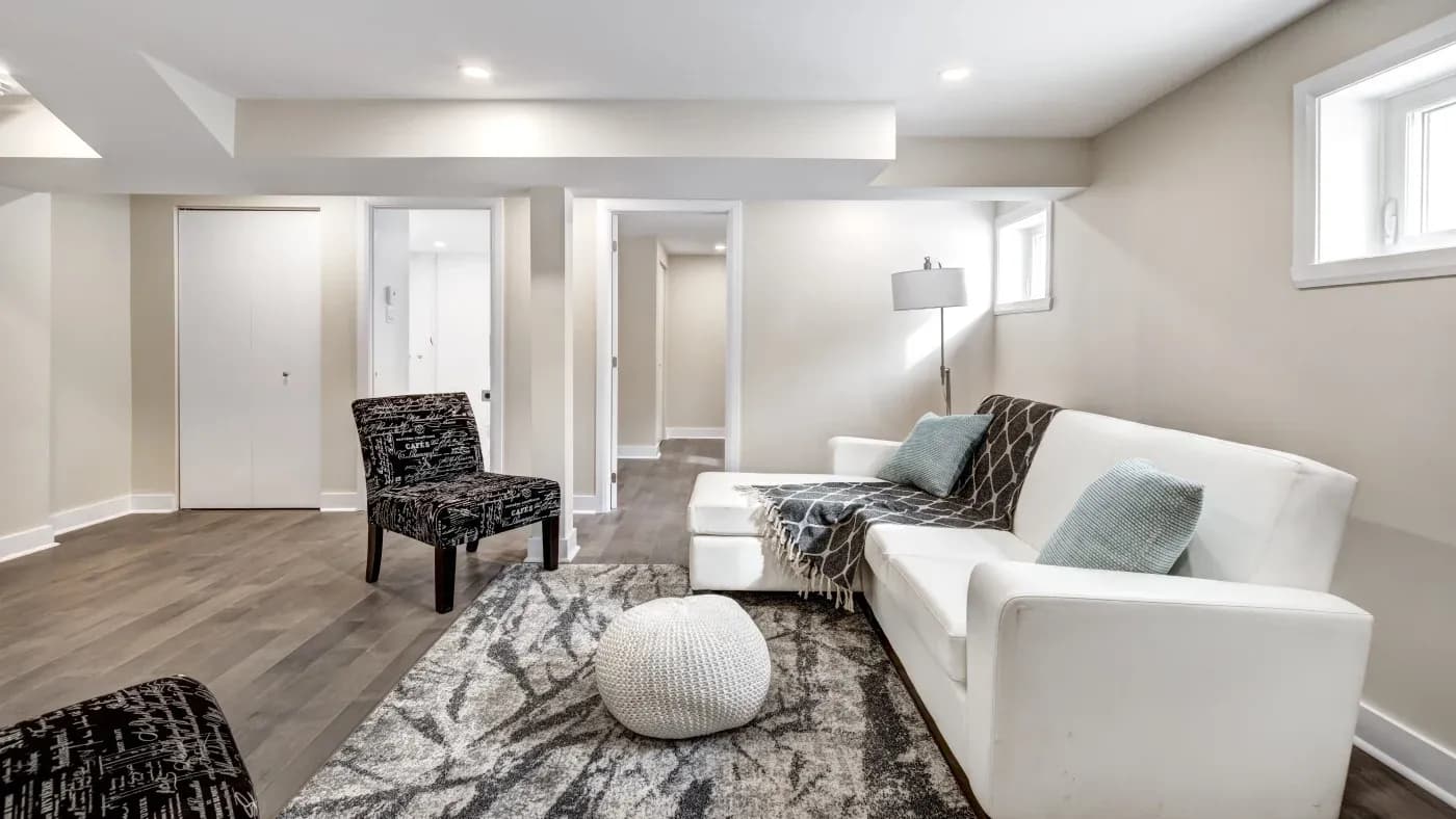 Modern living room with white sofa adorned with gray throw and blue pillows, black patterned chairs, and cozy gray carpet. Soft lighting and neutral walls.