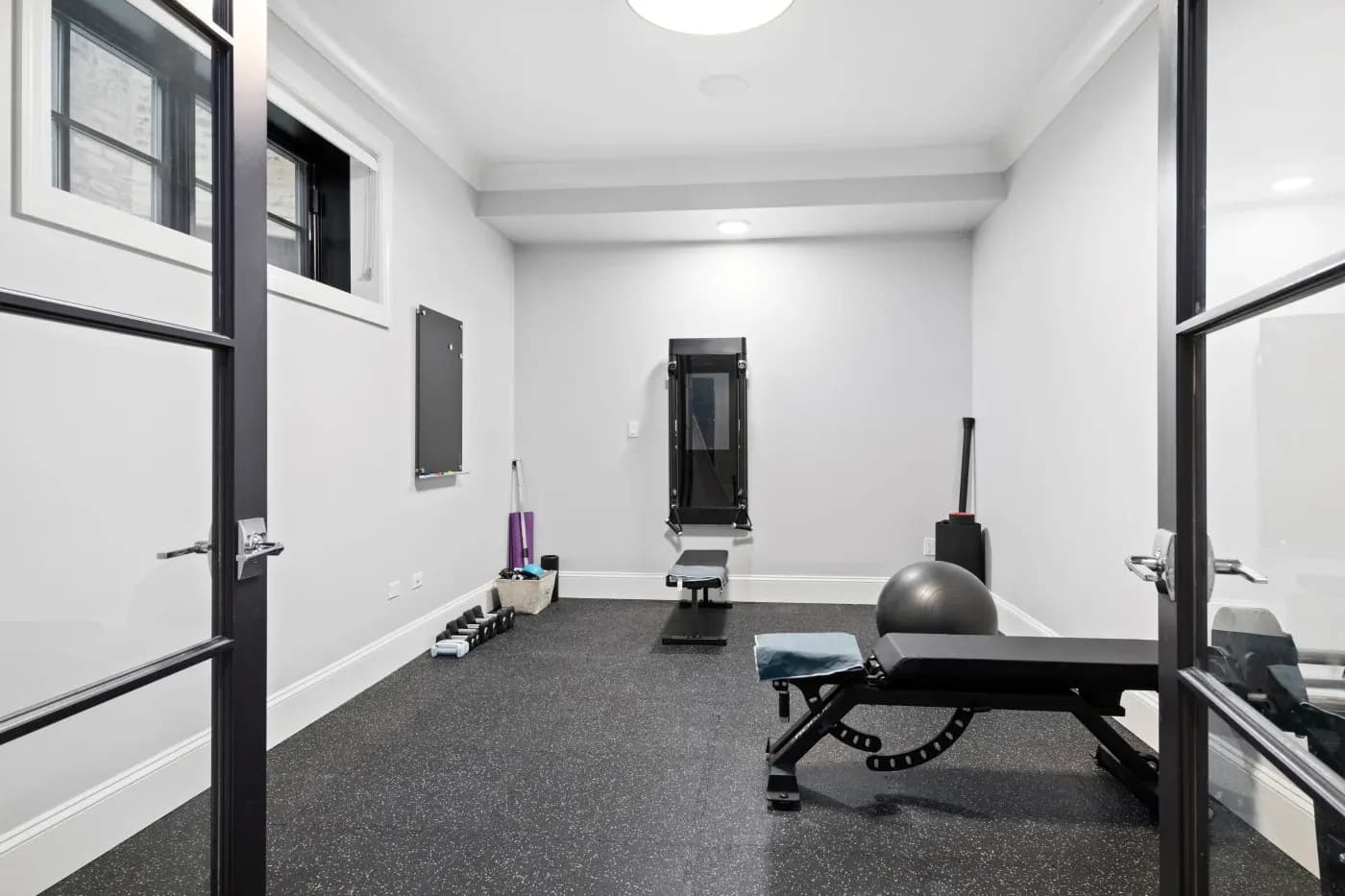 Home gym with black rubber flooring, featuring a bench, stability ball, wall-mounted exercise equipment, dumbbells, and a bright, open atmosphere.