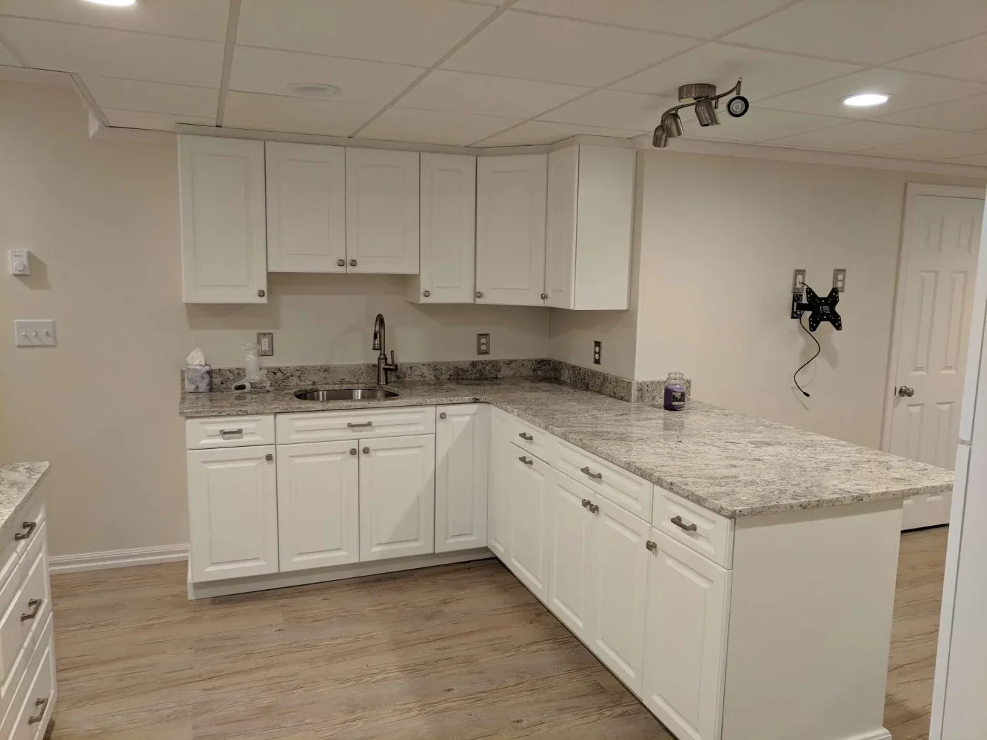 Clean white kitchen with L-shaped granite countertops, overhead cabinets, and a small sink. Simple decor, bright lighting, wood floor, and wall mount.