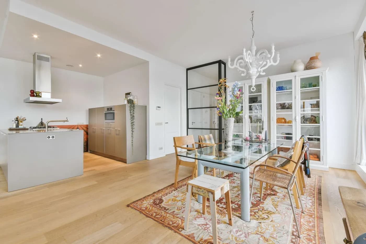Modern dining room with a glass table, woven chairs, and a floral rug. A chandelier hangs above. Adjacent is a sleek kitchen with gray cabinets. Light, airy feel.