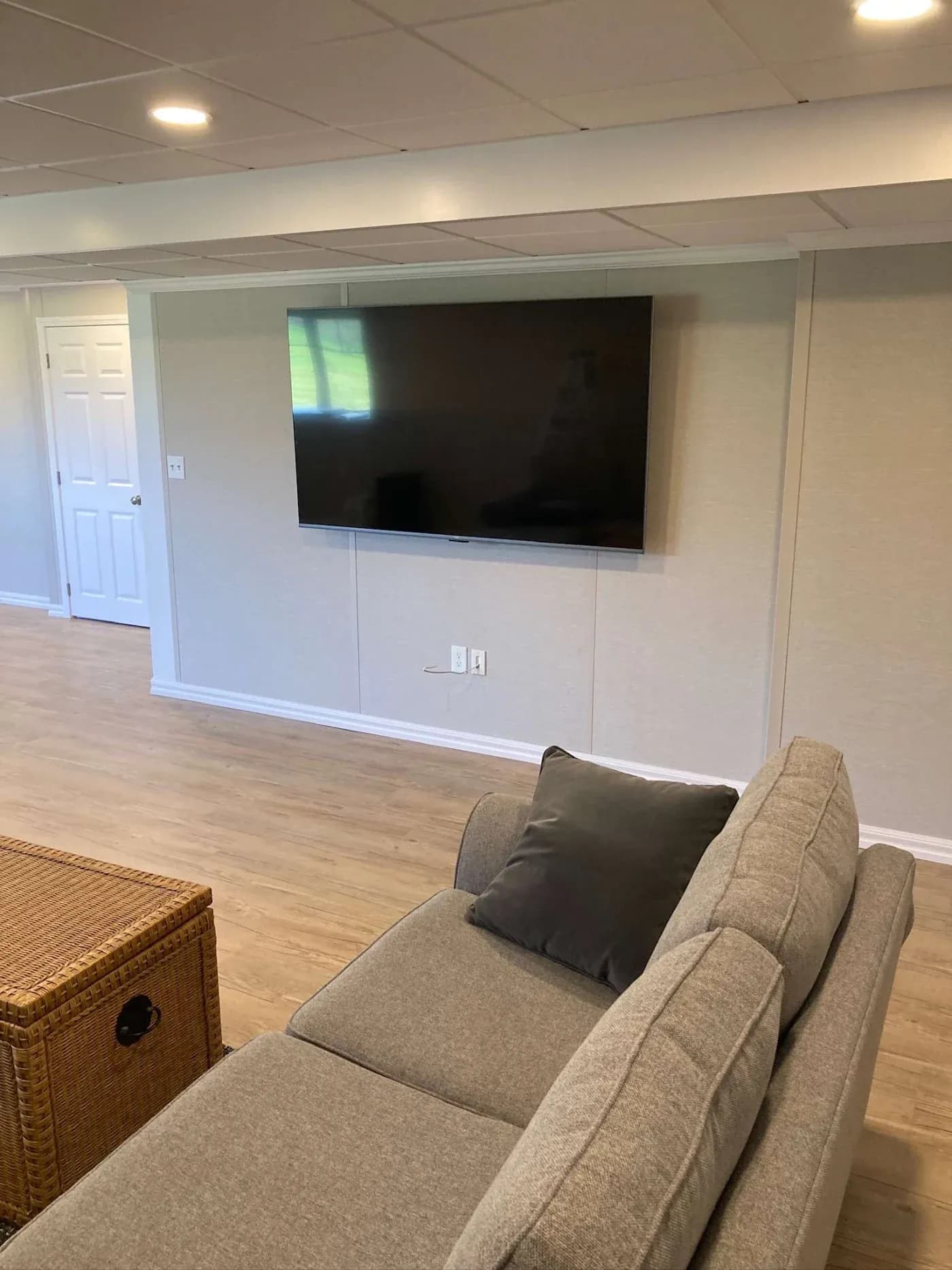 Modern basement with light wood flooring, a gray sectional sofa, wicker coffee table, and wall-mounted flat-screen TV, creating a cozy, inviting space.