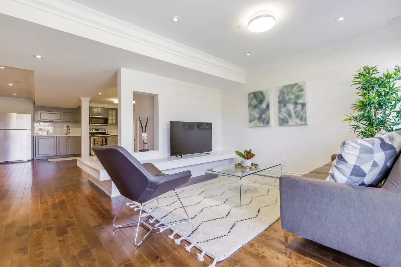Modern living room with a gray couch, black chair, and a glass coffee table on a patterned rug. Bright, minimal decor with plants and abstract art. Cozy atmosphere.
