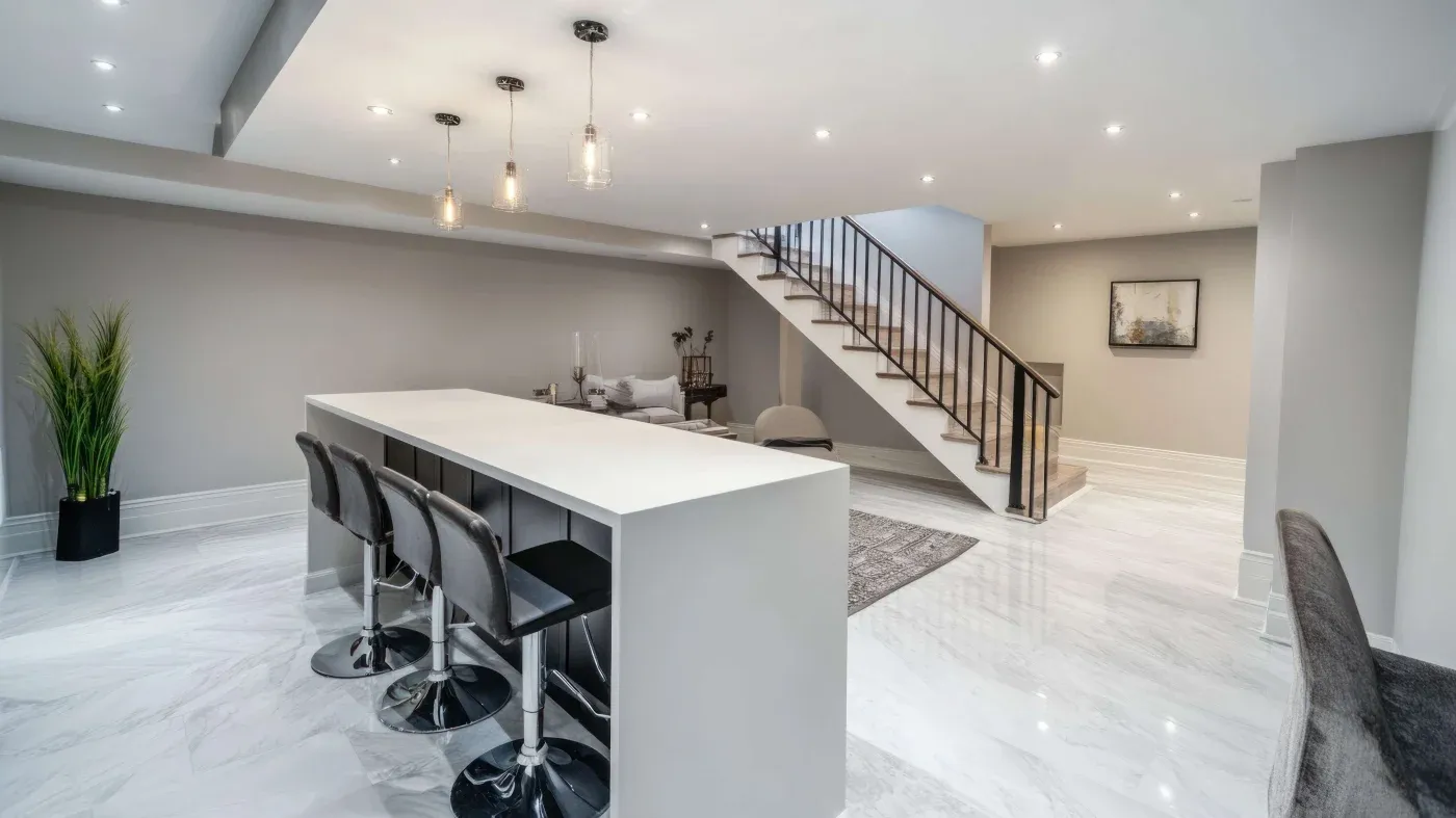 Spacious modern basement with sleek design. Features a long white bar with four stools, cozy gray seating area, staircase, and subtle lighting.