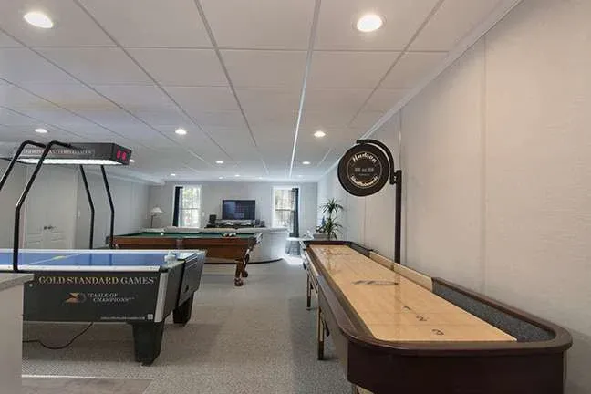 A spacious game room features an air hockey table, pool table, and shuffleboard. The room is well-lit with overhead lights and a cozy, inviting ambiance.