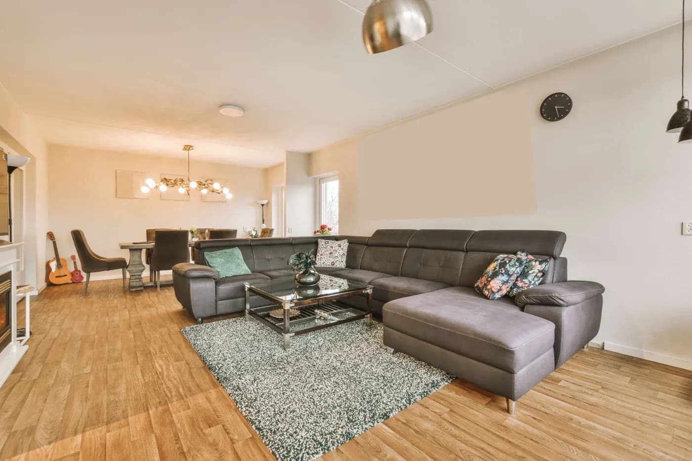 Modern living room with a large gray sectional sofa, colorful pillows, and a glass coffee table on a textured rug. Open dining area in background, wooden floor, contemporary decor, warm lighting.