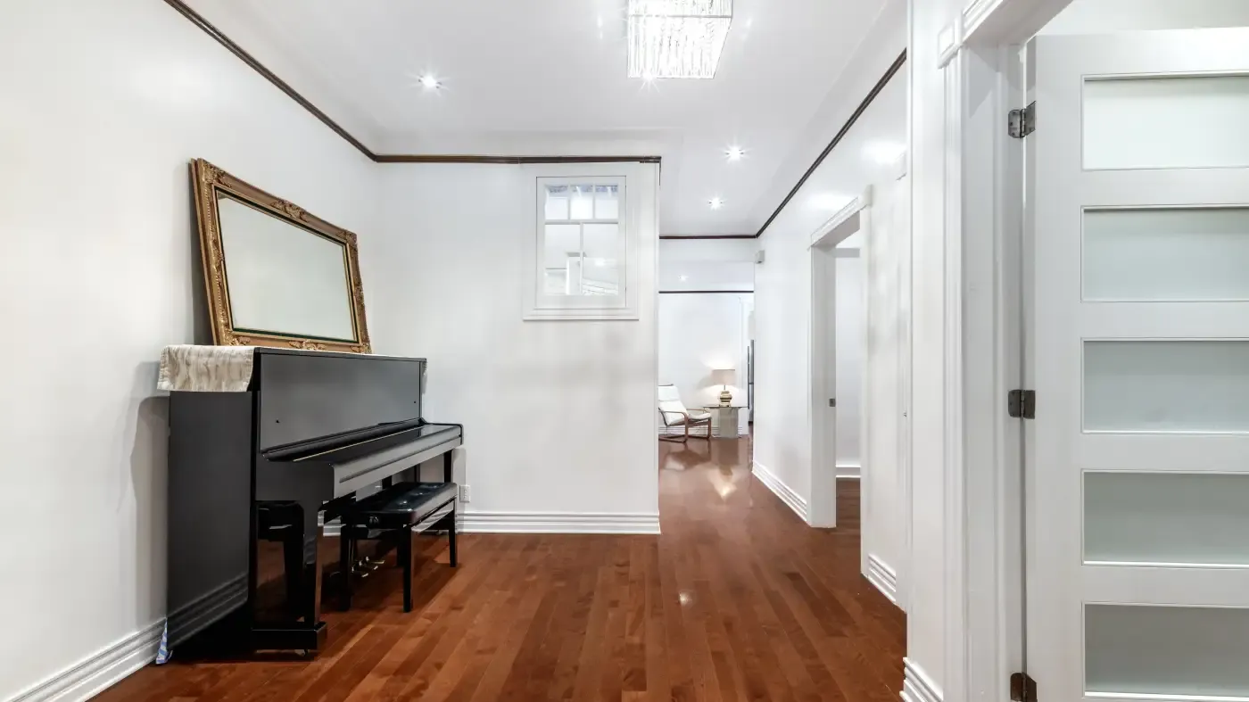 Bright hallway with wooden floors and white walls. A black piano with a gold-framed mirror sits on the left, leading into a room with modern furniture.