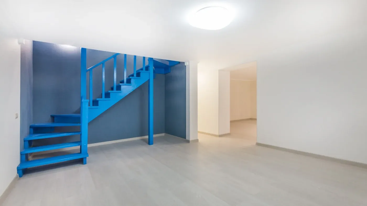 A minimalist room with light wood flooring features a vibrant blue staircase against a darker blue wall, leading to a bright, adjoining space.