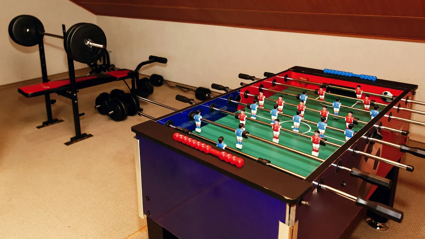 A foosball table and a weight bench with weights occupy a cozy attic room, conveying a relaxed recreational and fitness atmosphere.