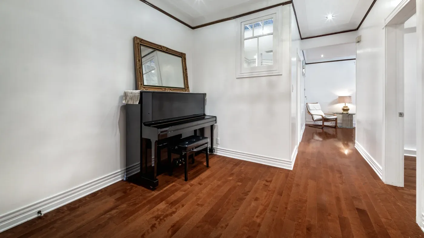 A hallway with warm wood flooring features an upright piano and mirror on the left. A cozy room with a chair and lamp is visible at the end. The atmosphere is inviting and elegant.