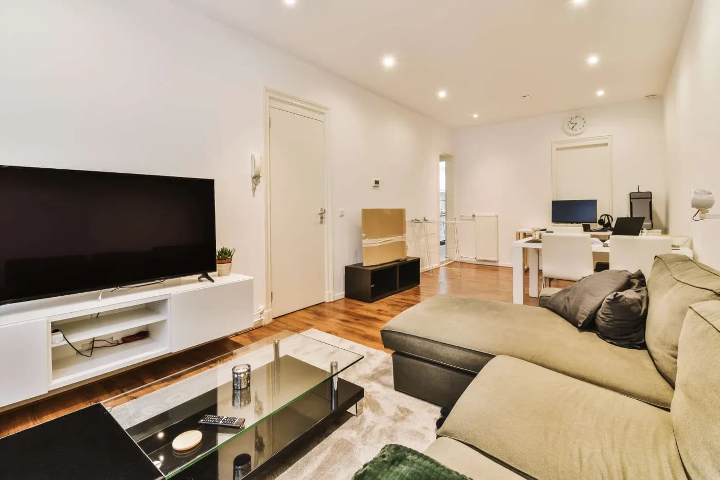 Modern living room with a large TV on a white stand, a cozy green sectional sofa, a glass coffee table, and a workspace in the background. Bright lighting.