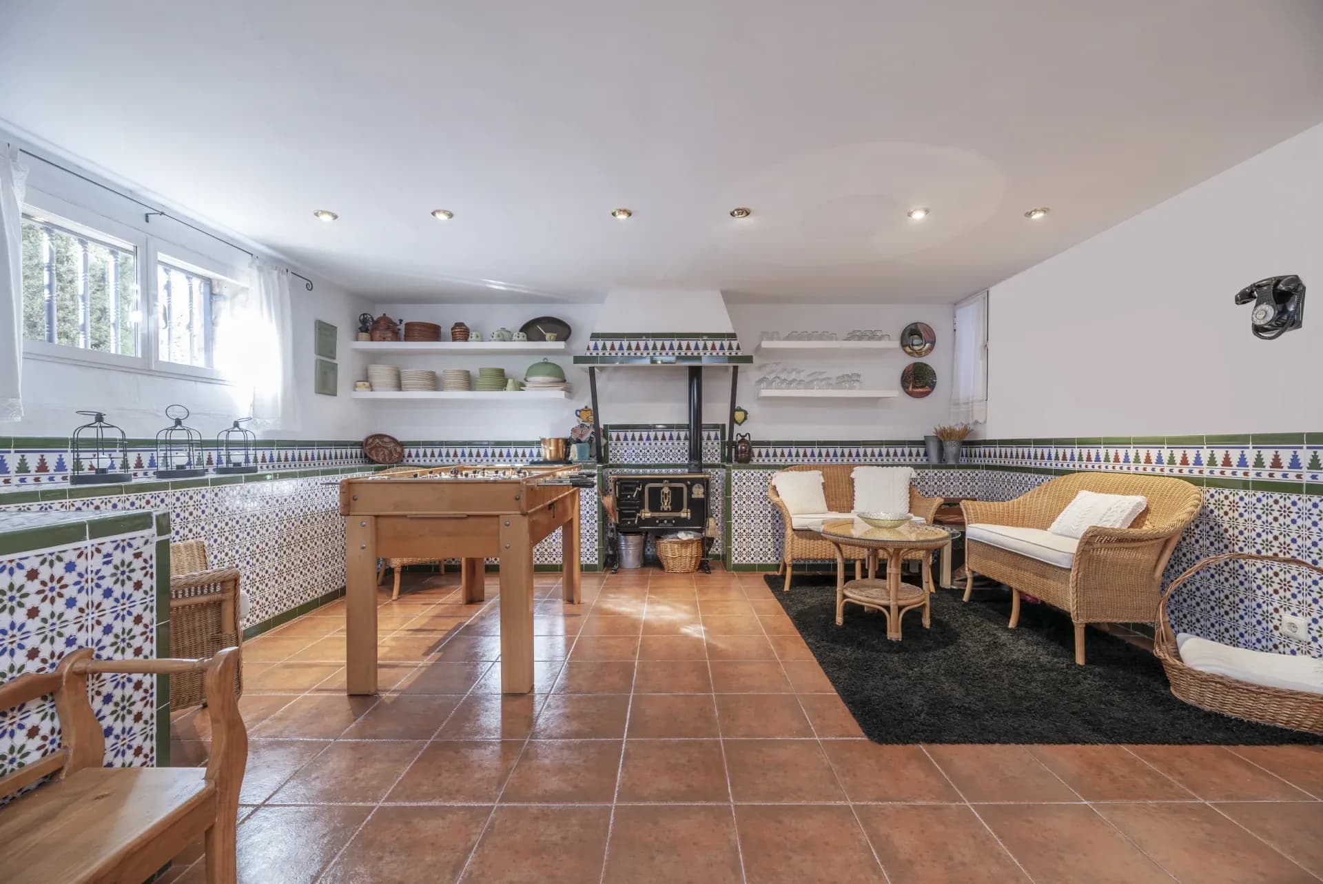 Spacious kitchen with colorful mosaic tiles, wooden table, wicker chairs, and a vintage stove. Bright and cozy ambiance with terracotta floor tiles.