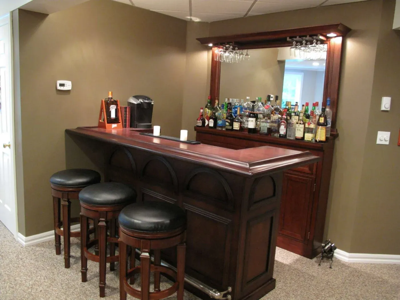 Cozy home bar with dark wood, three round black stools, and a wide mirror. Shelves display various liquor bottles, creating a warm, inviting atmosphere.