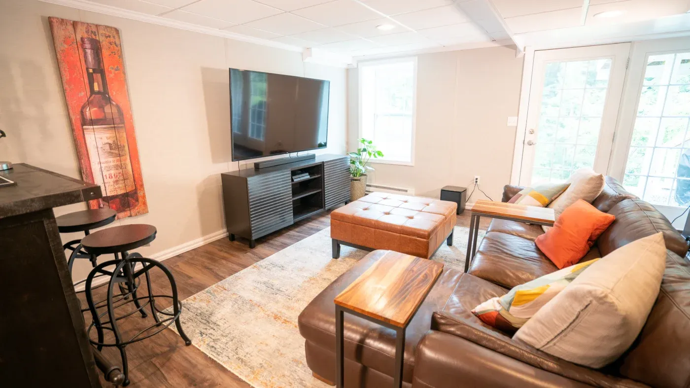 Cozy living room with a large TV, brown leather sectional, and orange accents. Features barstools, a wine-themed picture, and natural light.