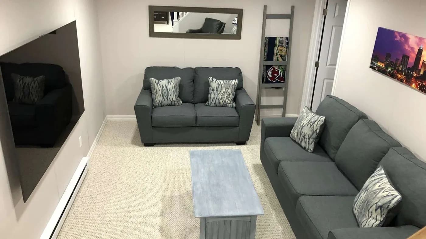 A cozy living room with gray sofas, patterned cushions, a light blue coffee table, and a flat-screen TV. A cityscape photo and mirror adorn the walls.