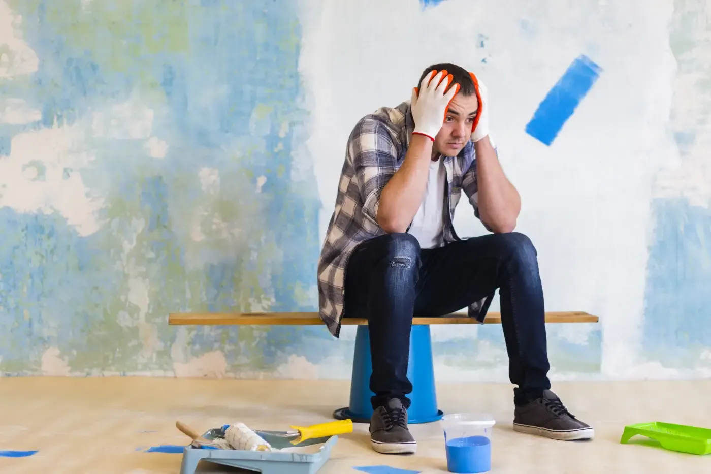 Man in a plaid shirt sits tired on a bench, surrounded by paint supplies in a room with blue and white walls. He appears frustrated and overwhelmed.