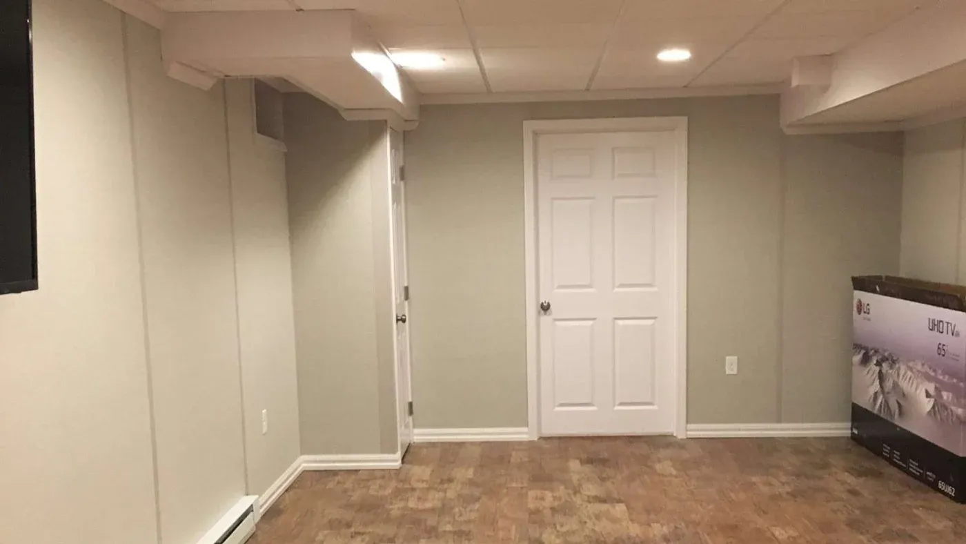 A plain basement room with beige walls, a white door, and a tiled floor. A boxed 65-inch LG UHD TV leans against the right wall, under soft lighting.