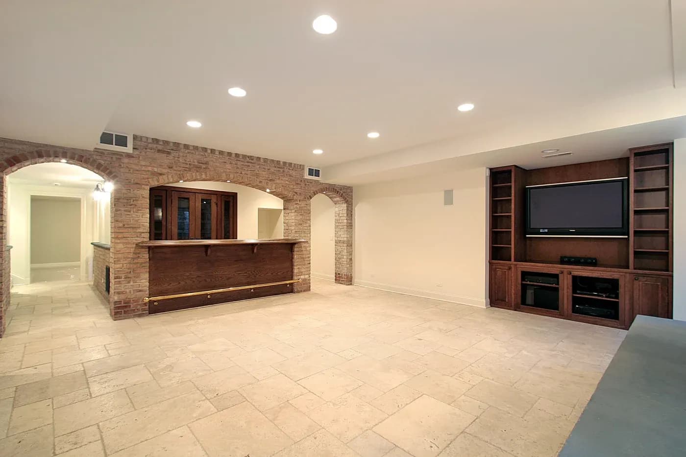 Spacious empty basement with beige tiles, brick archways, and a wooden bar. Wall-mounted TV with shelves, warm lighting, and a cozy ambiance.