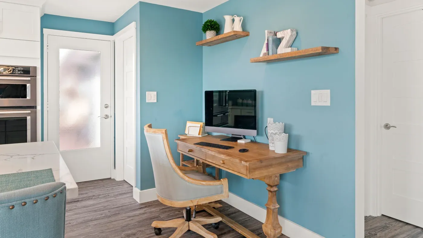 Cozy home office with a wooden desk and chair against a light blue wall. Shelves hold decor. A computer and framed picture add functionality. Calm ambiance.
