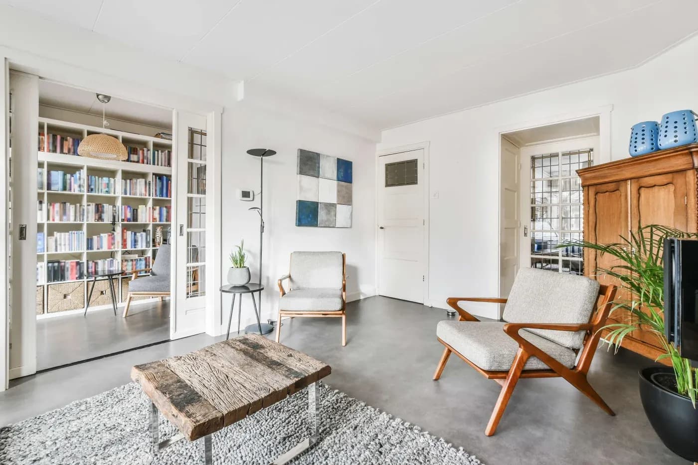 Bright living room with two mid-century modern chairs, a rustic wooden coffee table on a gray rug, and a view into a book-filled study. Cozy and inviting ambiance.