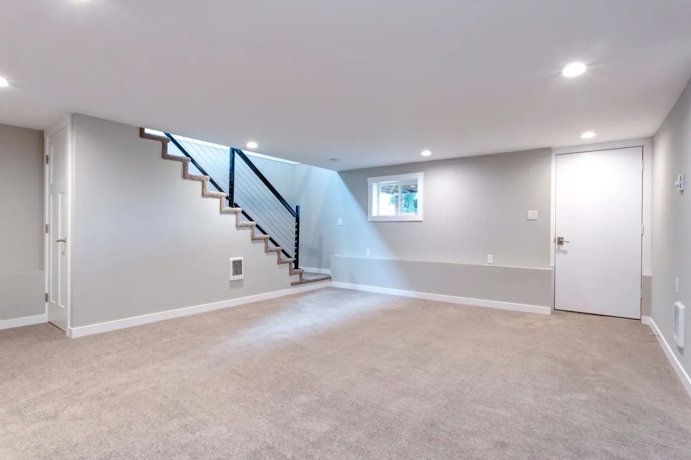 A modern, unfurnished basement room with beige carpeting and soft gray walls. It features recessed lighting, a white door, a small window, and a staircase with a sleek black railing, creating a clean and spacious atmosphere.