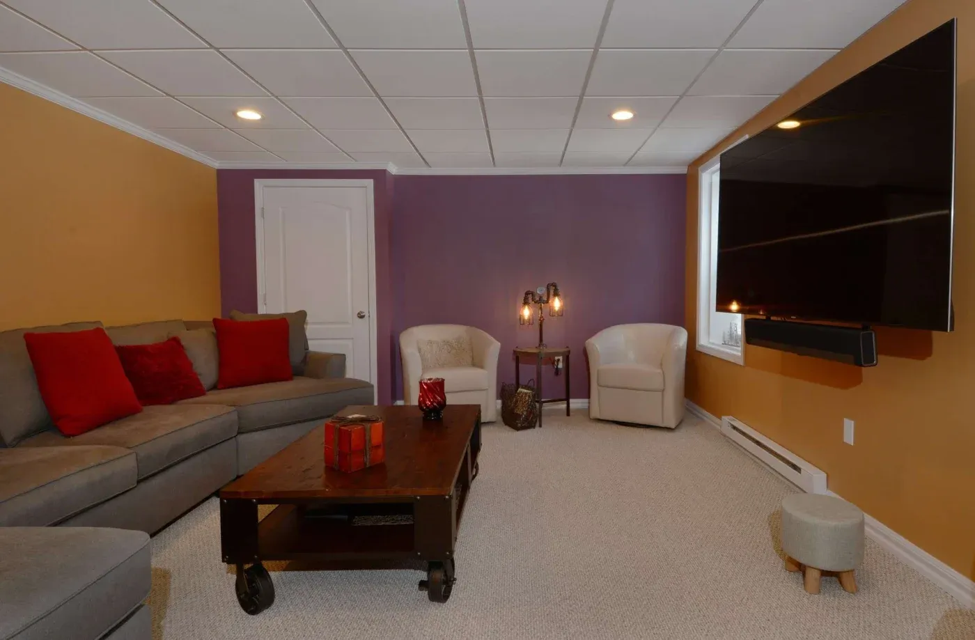 Cozy living room with a large gray sectional sofa and red pillows on the left, a wall-mounted TV on the right, and two white armchairs near a purple wall.