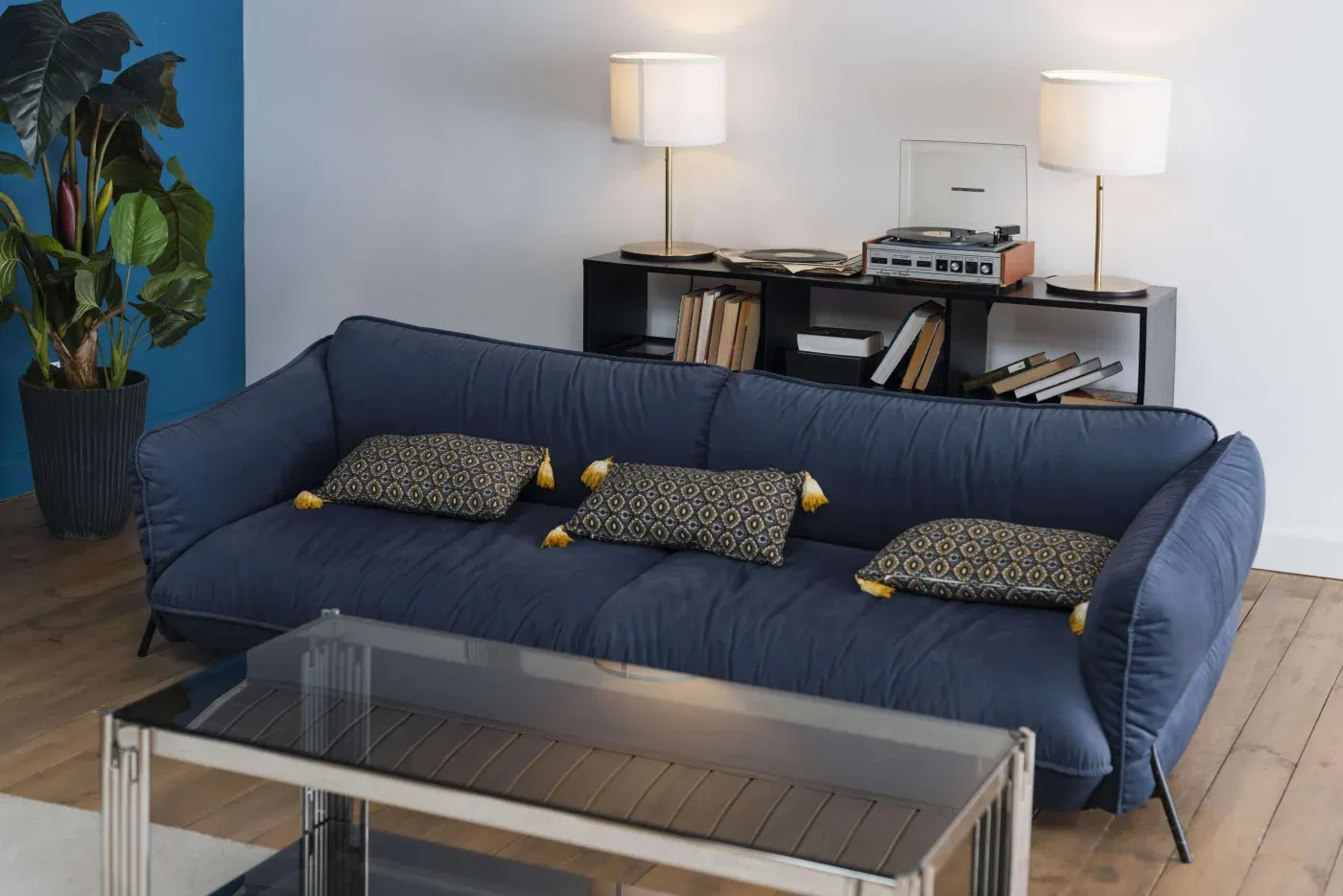 Cozy living room with a deep blue sofa, patterned cushions, and a glass coffee table. Bookshelf with lamps and a record player adds a retro touch.