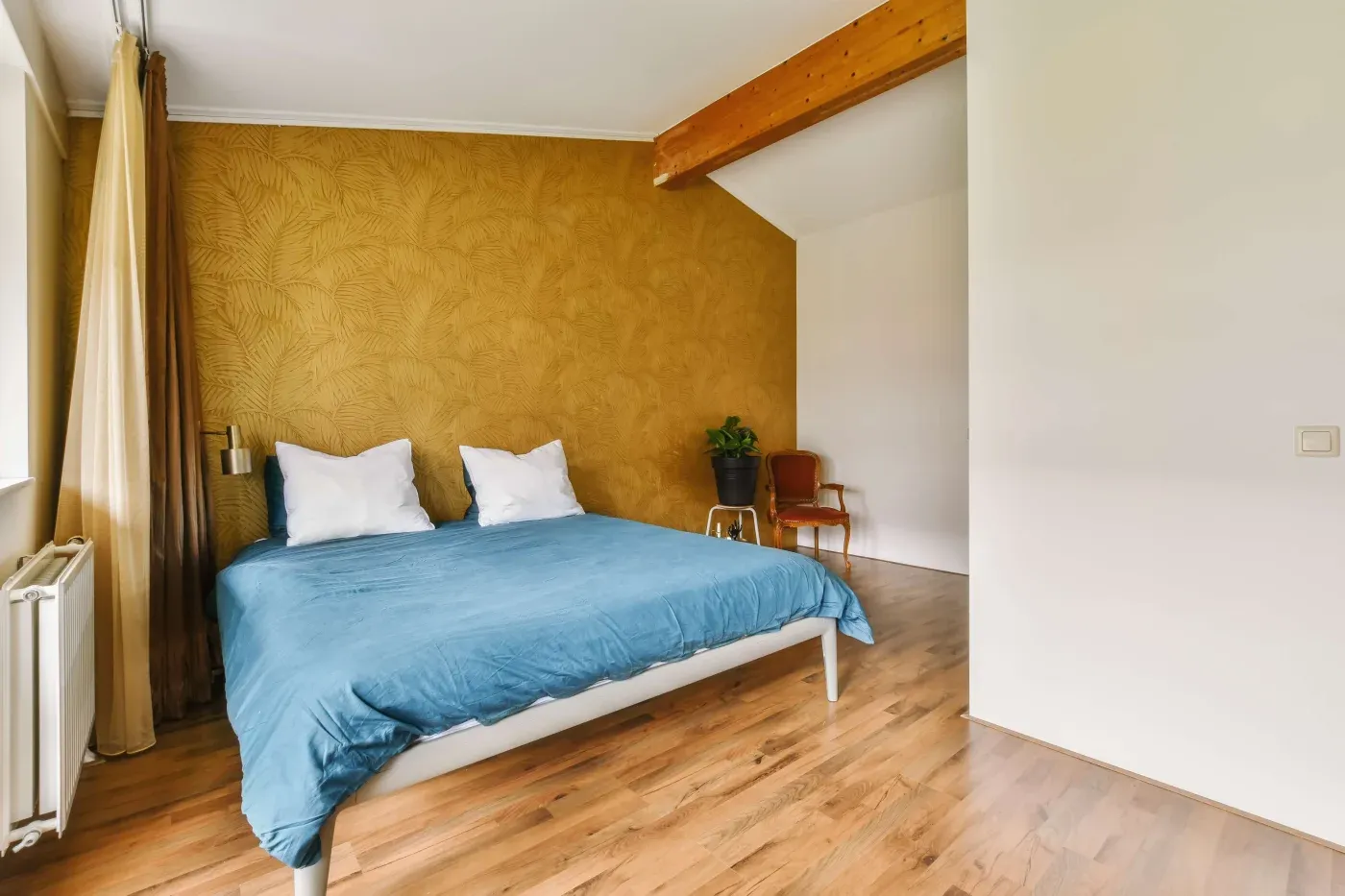 Cozy bedroom with a blue comforter, white pillows, wooden floor, and yellow textured wall. A potted plant and wooden chair add warmth. Natural light filters in.